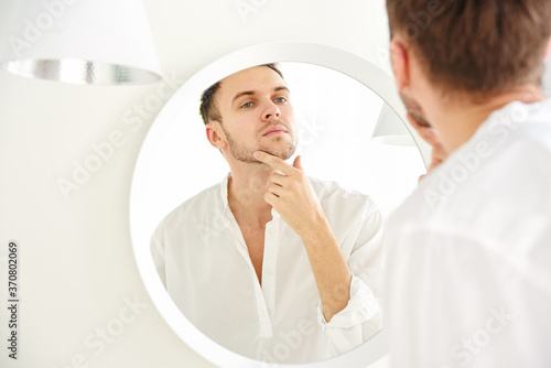 Young attractive Caucasian man examines his face in the mirror close-up. Skin care concept