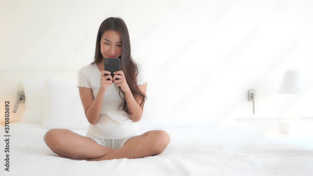 Attractive Asian woman in short white pajama set sitting on bed holding phone texting or playing game. Lady in bedroom smiling while using smartphone app or texting. White linen and sunlight. Attractive Asian woman in short white pajama set sitting on bed holding phone texting or playing game. Lady in bedroom smiling while using smartphone app or texting. White linen and sunlight.