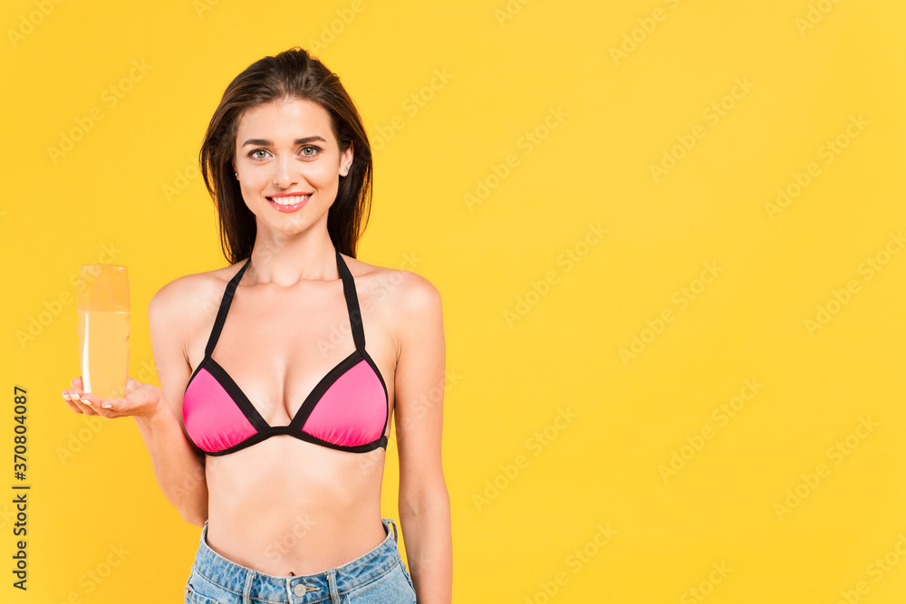  girl in swimsuit holding bottle with suntan isolated on yellow - obrazy, fototapety, plakaty 
