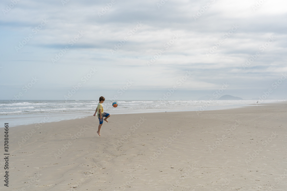 Menino, brasileiro, usando camiseta e bermuda, jugando futebol sozinho na praia de Guaratuba, litoral norte de São 
Paulo. 