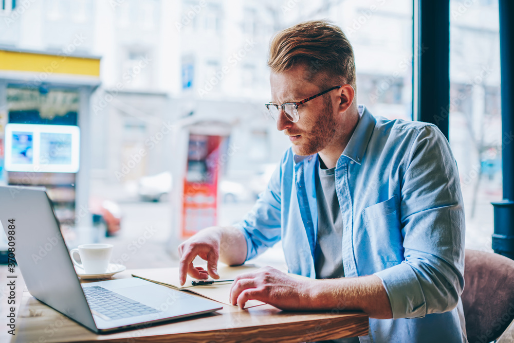 Fototapeta premium Concentrated hipster student reading information and checking data working at modern computer using wireless internet connection in coffee shop.Pensive young man preparing for exam at netbook