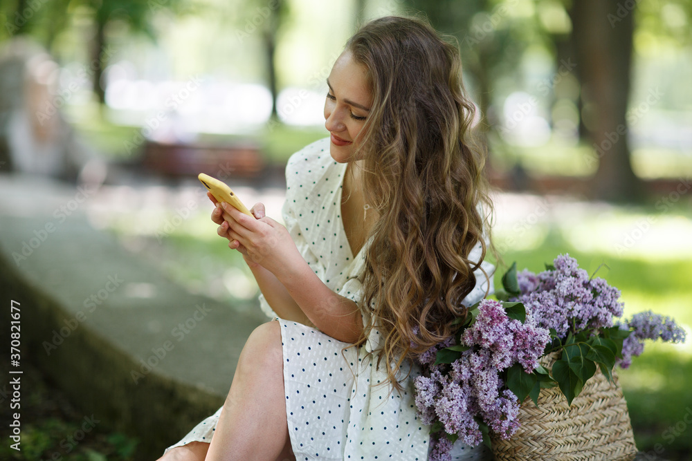 The girl in white dress holds in her hands a wicker basket with flowers and phone. Basket with lilacs. Girl and flowers. Sit with a basket of lilacs in the hands. Floristics The girl in white dress holds in her hands a wicker basket with flowers and phone. Basket with lilacs. Girl and flowers. Sit with a basket of lilacs in the hands. Floristics