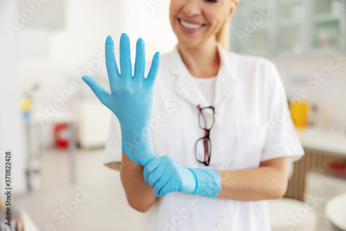 Closeup of smiling blond lab assistant standing in laboratory and putting rubber gloves on hands.