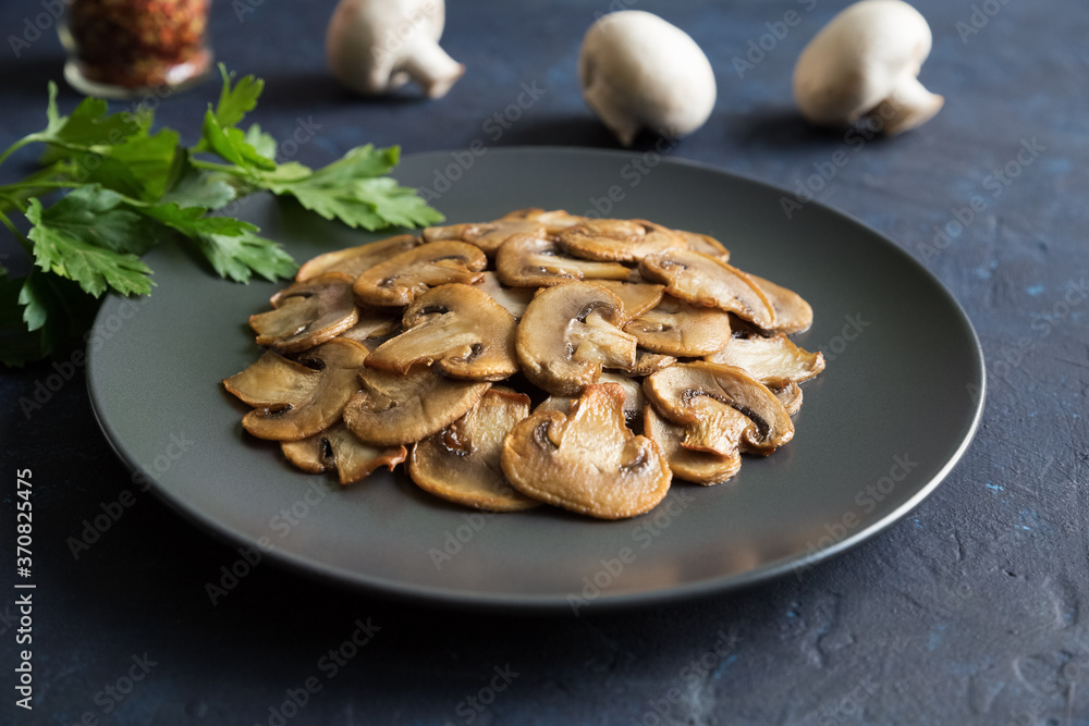 Fototapeta premium Slices of fried mushrooms on a black plate with of parsley leaves and several whole champignons. Close up on a dark blue background.
