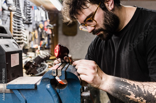 Side view of concentrated male mechanic with tattoos using vise and repairing red back lamp of motorbike while working in workshop photo