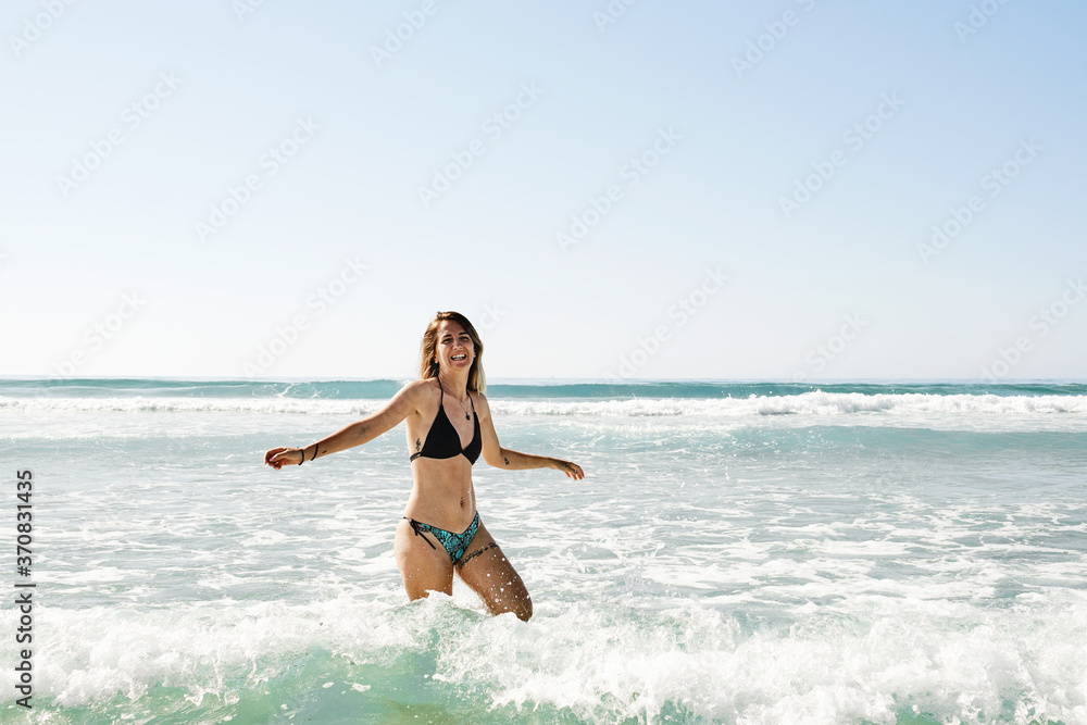 Cheerful female tourist wearing bikini walking in water along seashore and enjoying vacation looking at camera