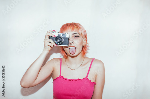 Naughty female millennial with piercing and pink hair taking pictures on vintage photo camera while showing tongue looking at camera on a white wall background photo