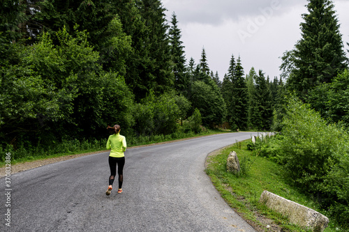 A young woman running outdoors. A girl in trendy long sleeve t-shirt is jogging on the road surrounded pines forest © Vadim Pastuh