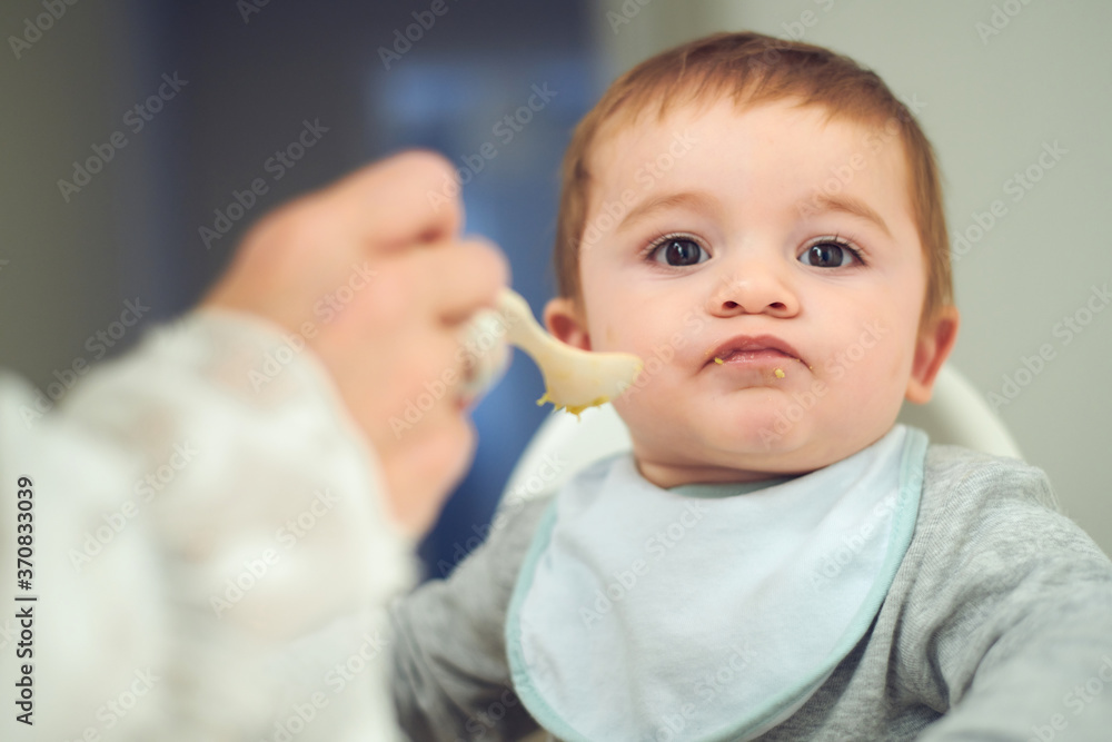 Adorable little baby sitting on high chair and eating fresh avocado from spoon held by crop anonymous mother Adorable little baby sitting on high chair and eating fresh avocado from spoon held by crop anonymous mother