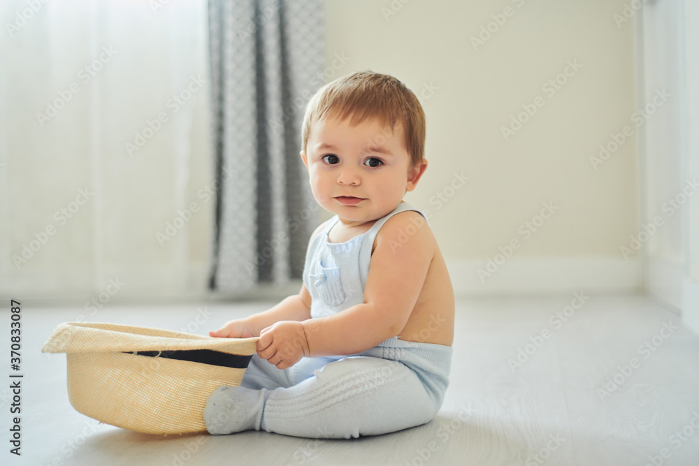 Adorable little baby holding big hat and looking at camera while sitting on floor and playing in light nursery at home Adorable little baby holding big hat and looking at camera while sitting on floor and playing in light nursery at home