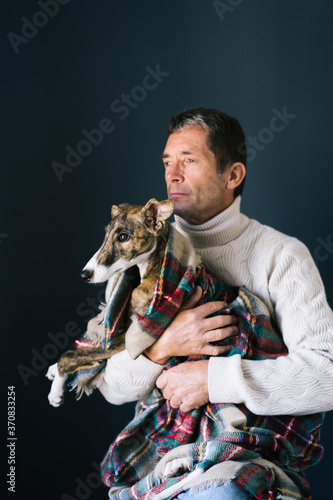 Adult man in sweater looking away and embracing adorable Whippet dog in checkered blanket while sitting against gray background photo