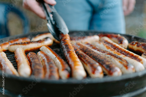 Crop anonymous person in jeans frying delicious beef sausages on grill and using tongs while having barbecue picnic on backyard photo