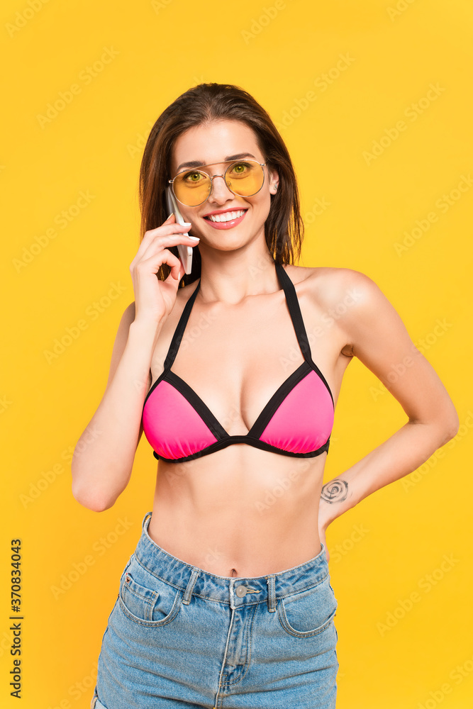  girl in swimsuit and sunglasses talking on smartphone while standing with hand on hip isolated on yellow - obrazy, fototapety, plakaty 