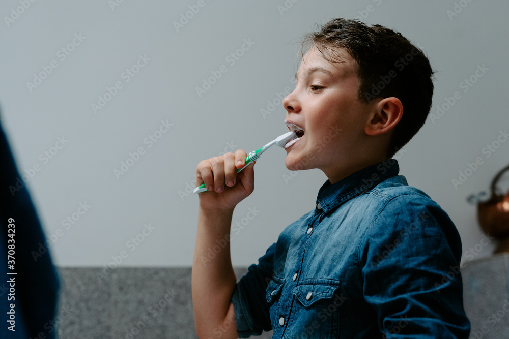 Side view adorable positive boy in denim shirt brushing teeth with plastic toothbrush in modern bathroom after waking up in morning Side view adorable positive boy in denim shirt brushing teeth with plastic toothbrush in modern bathroom after waking up in morning