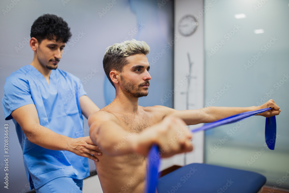 Sports medicine doctor checking up back of sportive man with dyed hair stretching elastic tape during rehabilitation period and looking away Sports medicine doctor checking up back of sportive man with dyed hair stretching elastic tape during rehabilitation period and looking away