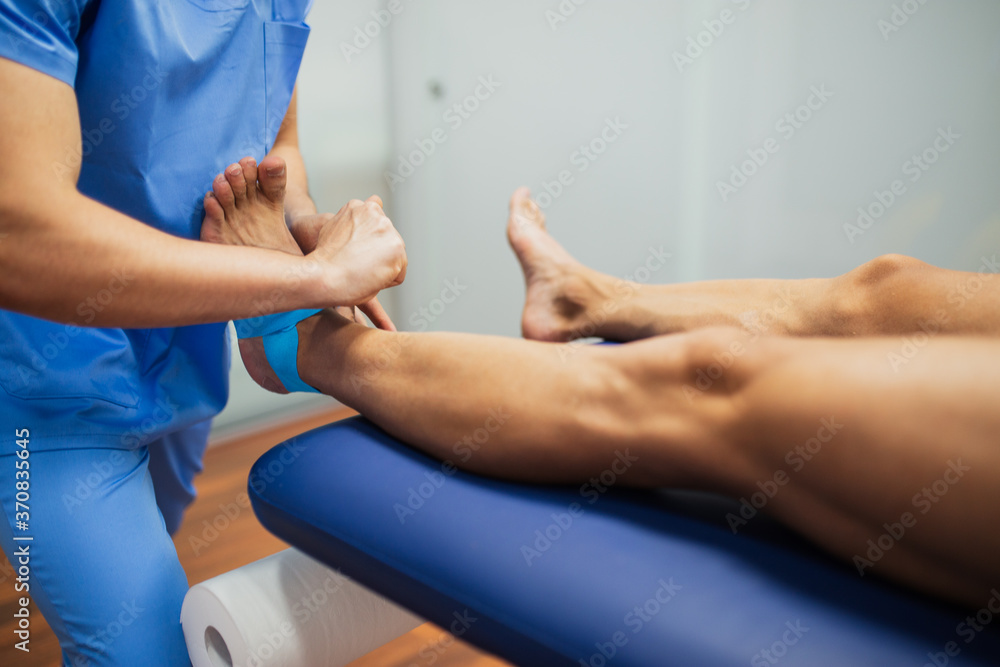 Top view of crop unrecognizable chiropractor in blue uniform attaching kinesio tape on foot of faceless man lying on examination couch in clinic Top view of crop unrecognizable chiropractor in blue uniform attaching kinesio tape on foot of faceless man lying on examination couch in clinic