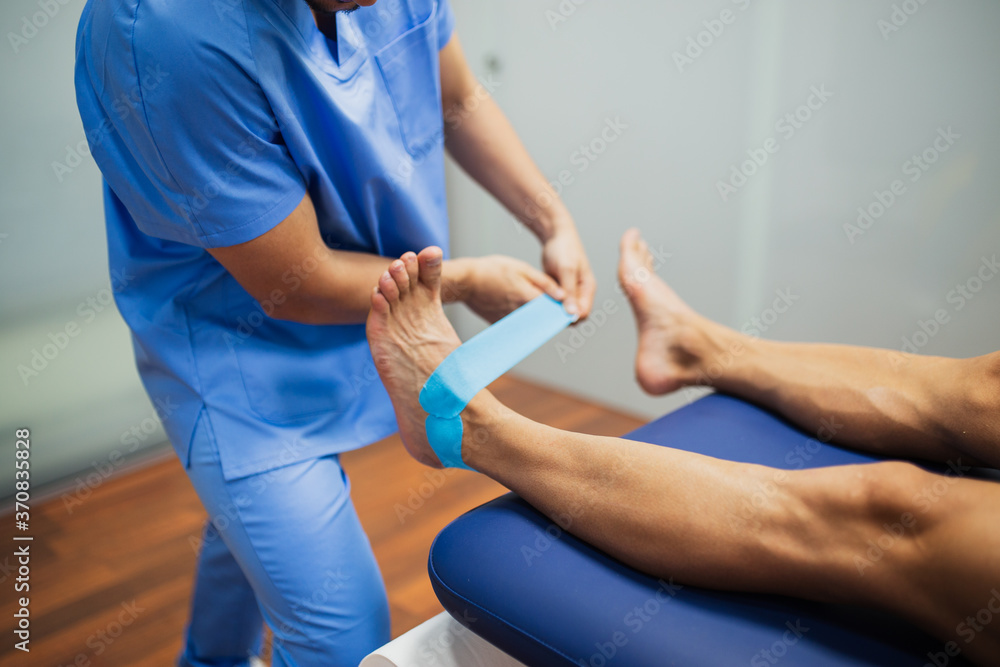 Top view of crop unrecognizable chiropractor in blue uniform attaching kinesio tape on foot of faceless man lying on examination couch in clinic Top view of crop unrecognizable chiropractor in blue uniform attaching kinesio tape on foot of faceless man lying on examination couch in clinic