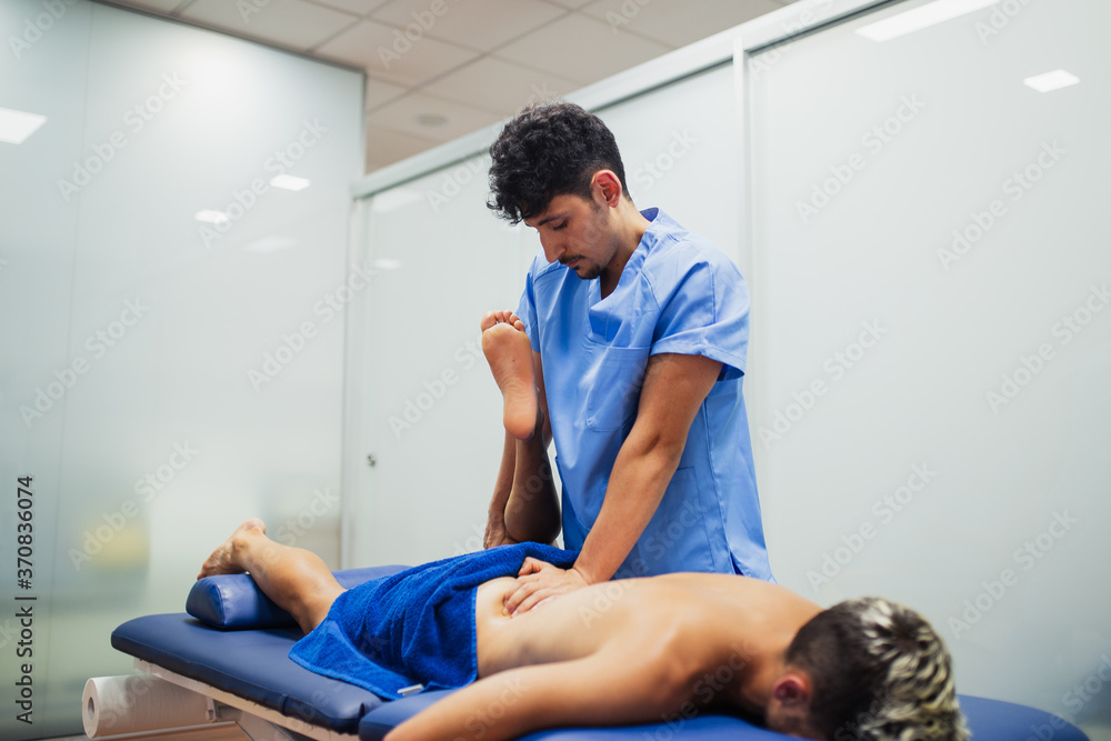 Osteopath in blue uniform examining leg of unrecognizable slim male patient with dyed hair lying on examination table in clinic Osteopath in blue uniform examining leg of unrecognizable slim male patient with dyed hair lying on examination table in clinic