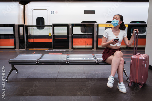 Young concentrated Asian lady wearing mini skirt and face mask browsing mobile phone while sitting on bench with suitcase in train station photo