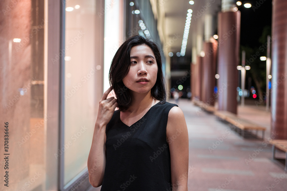 Ethnic female standing on sidewalk in evening near illuminated building and looking away Ethnic female standing on sidewalk in evening near illuminated building and looking away