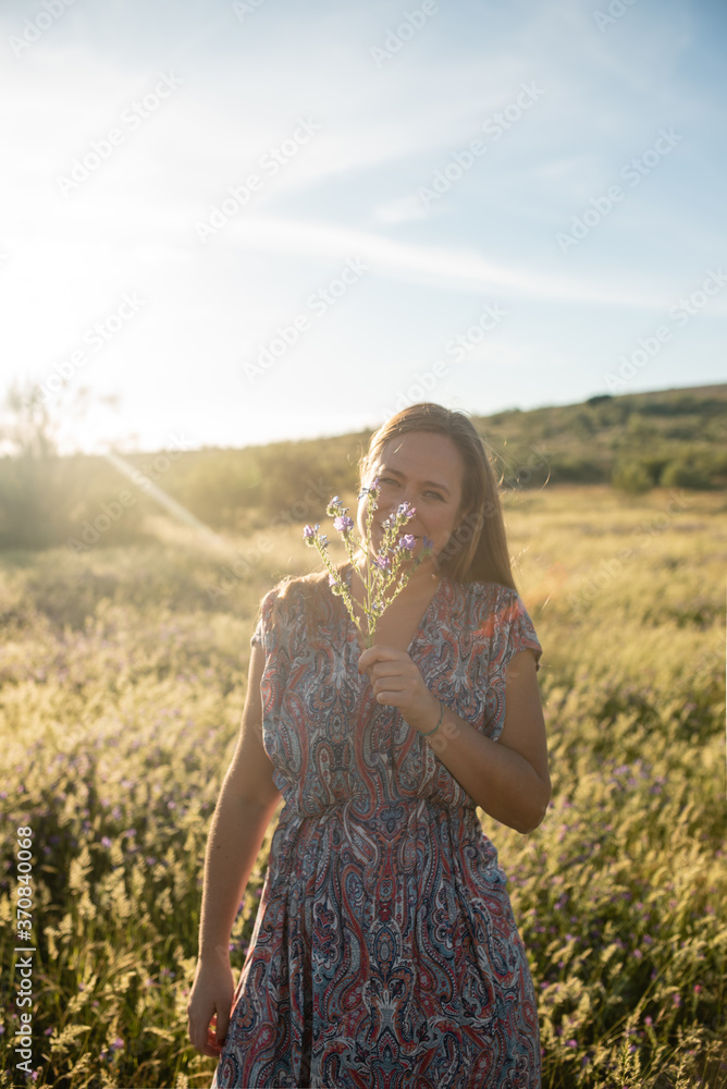 Smiling female wearing trendy dress and straw hat standing on meadow during sunny day and enjoying summer holding fresh perfumed flower Smiling female wearing trendy dress and straw hat standing on meadow during sunny day and enjoying summer holding fresh perfumed flower