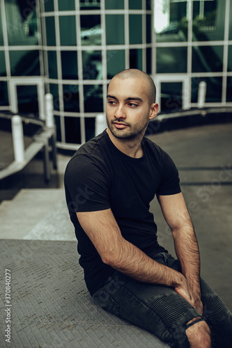 Modern young bearded hipster guy in casual black shirt and ripped jeans looking away while sitting on stairs near contemporary building