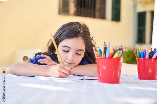 Positive school aged girl while sitting at table and drawing with colorful pencils in copybook during free time photo