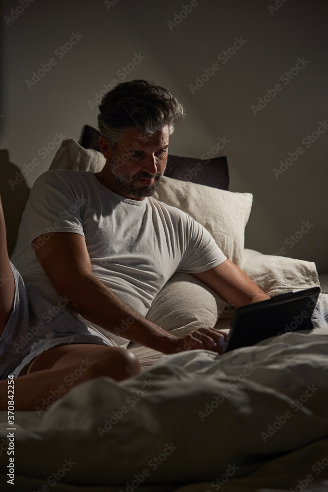 Smiling middle aged bearded male in casual clothes lying on bed and browsing laptop while resting in cozy bedroom in evening Smiling middle aged bearded male in casual clothes lying on bed and browsing laptop while resting in cozy bedroom in evening