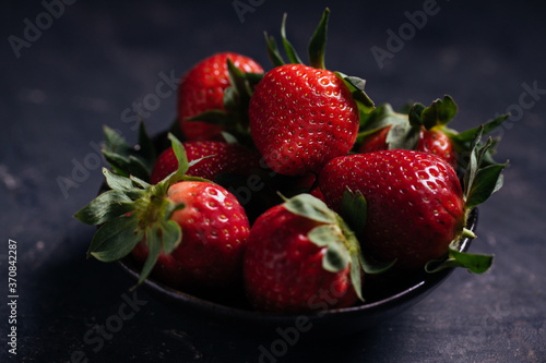Closeup of delectable juicy ripe strawberries with green leaves arranged in black ceramic bowl on dark background
