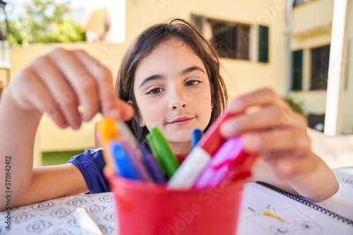 Positive school aged girl while sitting at table and choose drawing with colorful pencils in copybook during free time