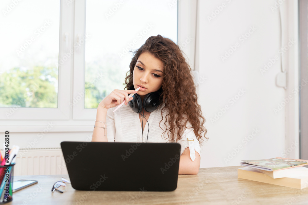 Beautiful girl with curly hair wearing headphones around neck, holding pen and looking at laptop by the window. Beautiful girl with curly hair wearing headphones around neck, holding pen and looking at laptop by the window.