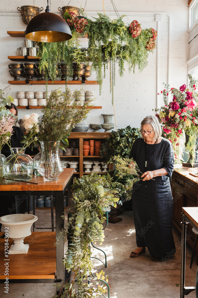 Professional adult female florist choosing green decorative plants for composition while working with colleague in cozy floristry studio Professional adult female florist choosing green decorative plants for composition while working with colleague in cozy floristry studio