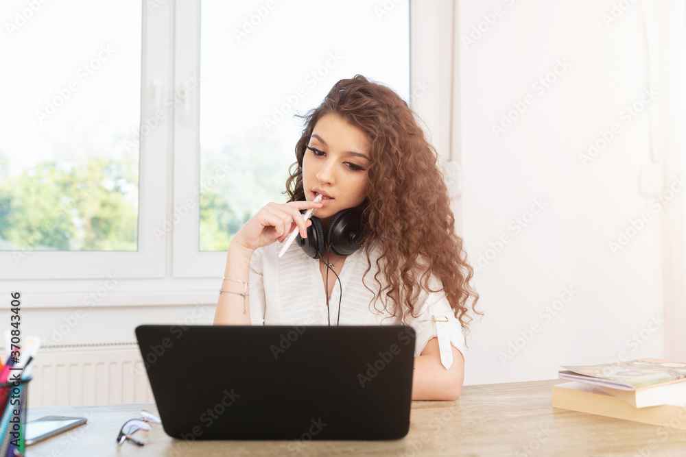 Cute brunette sitting in office by the window wearing headphones around neck, looking at laptop and holding a pen. Cute brunette sitting in office by the window wearing headphones around neck, looking at laptop and holding a pen.