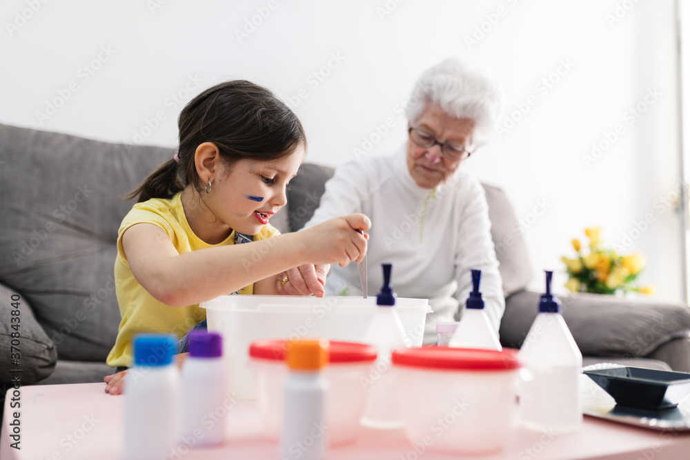 Smiling granny in eyeglasses helping positive granddaughter with dirty face in casual wear preparing blend using colorants in plastic container while sitting on comfortable couch in living room Smiling granny in eyeglasses helping positive granddaughter with dirty face in casual wear preparing blend using colorants in plastic container while sitting on comfortable couch in living room