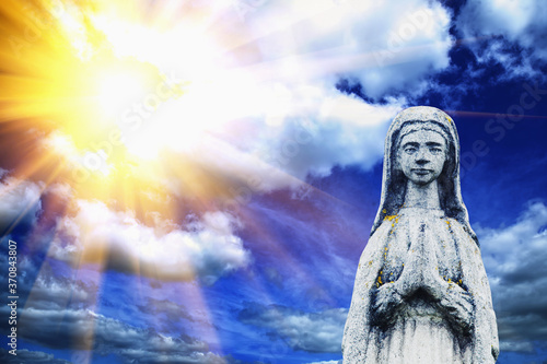 Virgin Mary against dramatic view of cloudy sky. An ancient stone statue. Prayer, faith, religion, love, hope concept.