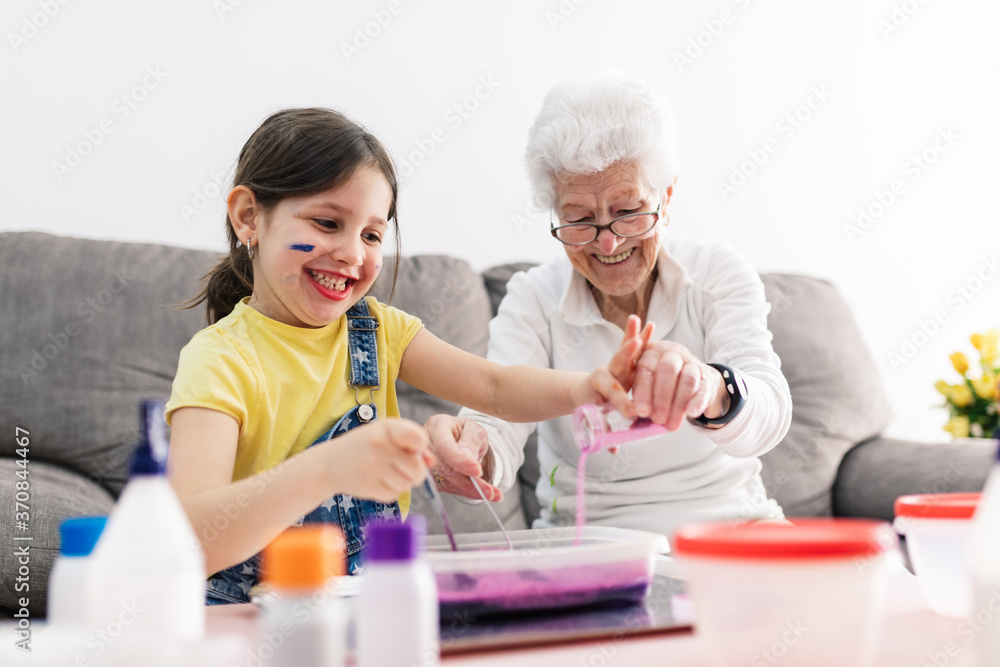 Smiling granny in eyeglasses helping positive granddaughter with dirty face in casual wear preparing blend using colorants in plastic container while sitting on comfortable couch in living room Smiling granny in eyeglasses helping positive granddaughter with dirty face in casual wear preparing blend using colorants in plastic container while sitting on comfortable couch in living room
