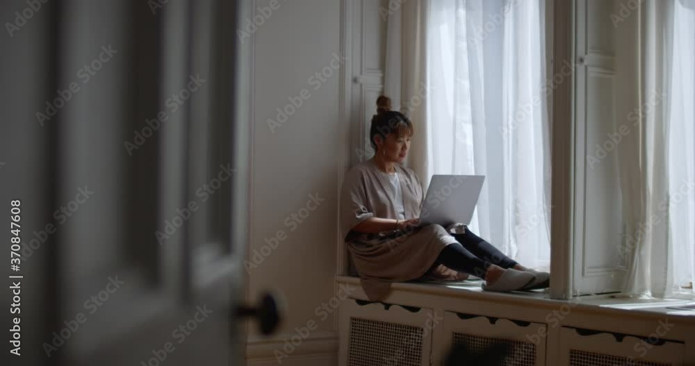 Warm sunlight peeks through window and sheer curtains as woman writes an email on her laptop while sitting on a window sill working from home Warm sunlight peeks through window and sheer curtains as woman writes an email on her laptop while sitting on a window sill working from home
