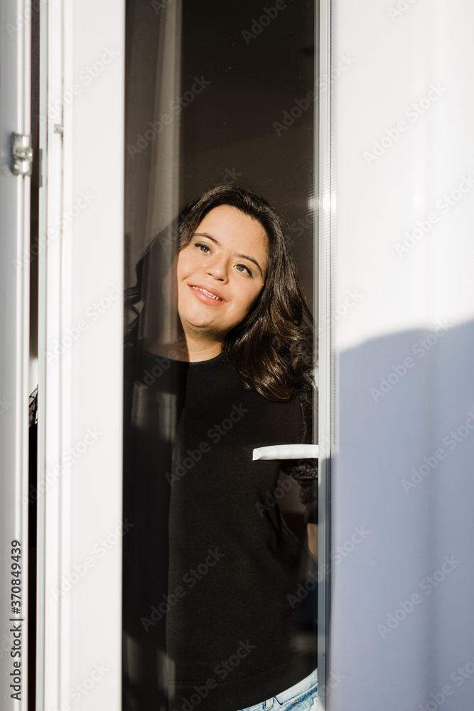 Thoughtful woman with down syndrome smiling while looking through window at home Thoughtful woman with down syndrome smiling while looking through window at home