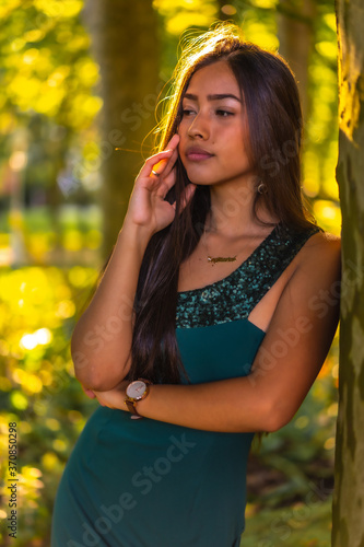 A young brunette Latina with long straight hair leaning against a tree in a green dress. Portrait with a sweet and sensual look  vertical photo