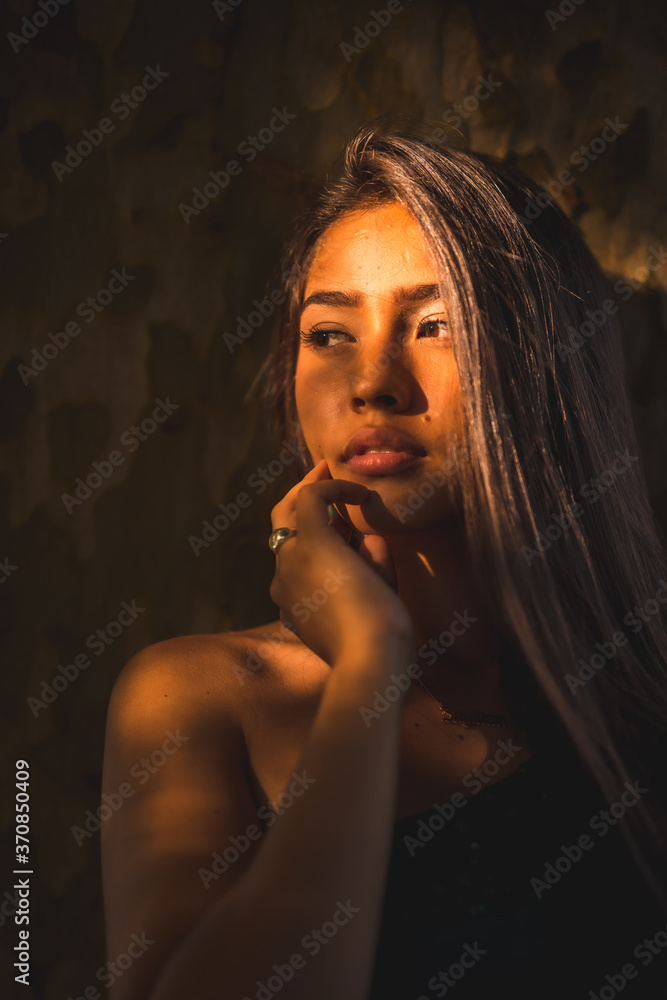A young Latina brunette leaning against a tree with a slight light on her face. Portrait with a sweet and sensual look A young Latina brunette leaning against a tree with a slight light on her face. Portrait with a sweet and sensual look