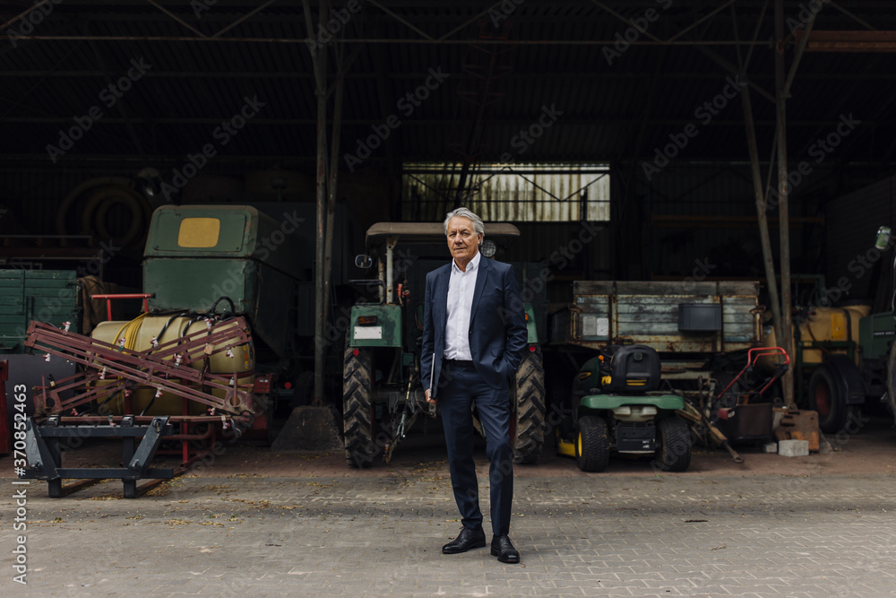 Senior businessman standing on a farm with tractor in barn Senior businessman standing on a farm with tractor in barn