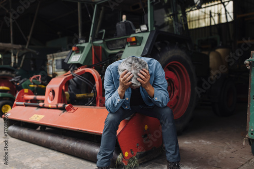 Despaired senior man on a farm sitting on tractor in barn