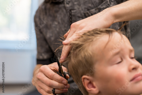 A pretty boy toddler happy to be on the haircut with a professional children's hairdresser. Blond little boy having a haircut at hair salon. Hairdresser's hands making hairstyle to child.