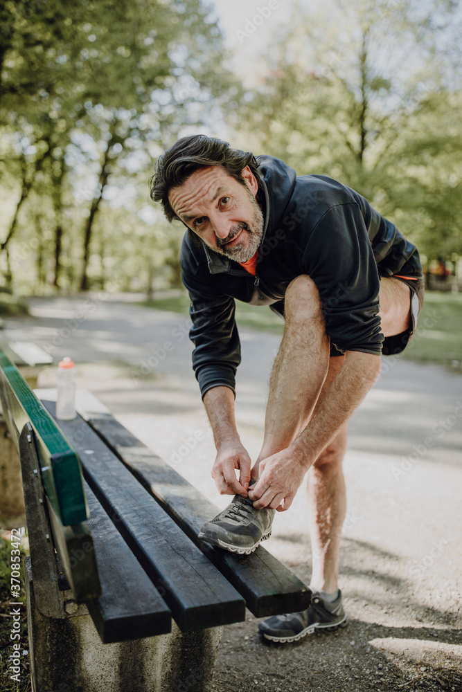 Smiling mature man tying shoelace on bench while standing in park Smiling mature man tying shoelace on bench while standing in park