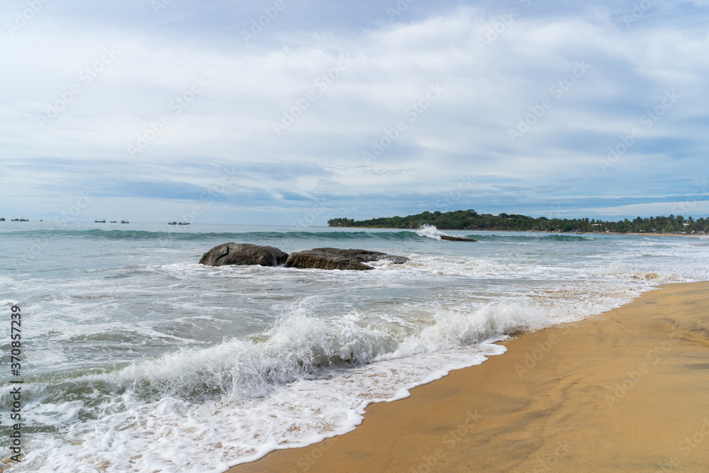 Tropical beach with rocks in the sea. palm trees on the background.  cloudy sky. Arugam bay, Sri Lanka