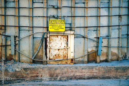 Close-up of a bright yellow caution sign hanging over a hatch at the base of an old concrete silo warning about a restricted space.