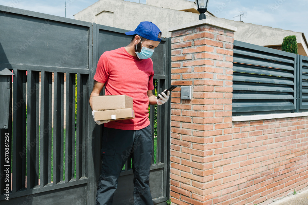 Male postal worker using mobile phone while standing with package against gate Male postal worker using mobile phone while standing with package against gate