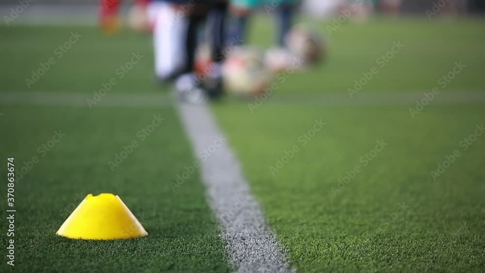 Yellow cone marker and soccer ball moving on green artificial turf with blurry kid soccer team training in soccer academy. Yellow cone marker and soccer ball moving on green artificial turf with blurry kid soccer team training in soccer academy.