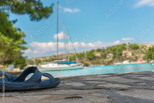 Closeup view a rubber slipper on the shore of the sea, blurred sailboats, hills and teal sea in the background.