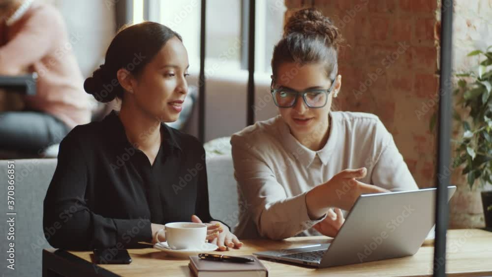 Two young multiethnic businesswomen sitting at table in cafe, discussing something on laptop and smiling while having coffee on break Two young multiethnic businesswomen sitting at table in cafe, discussing something on laptop and smiling while having coffee on break